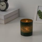 Lit candle in green glass holder beside books, clock, and framed plant. Provides warm, ambient light.