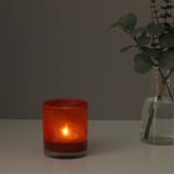 A small, red, glass candle holder glows softly on a table, casting a warm light. Beside it, a glass vase holds green leaves.