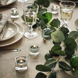 A festive table setting with green foliage, white plates, clear glasses, and small VINTERFINT green candles creating a cozy atmosphere.
