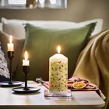 Festive decorated white candle, lit, on table; sofa, green pillows; orange slices nearby.