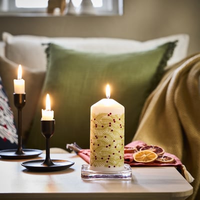 Festive decorated white candle, lit, on table; sofa, green pillows; orange slices nearby.