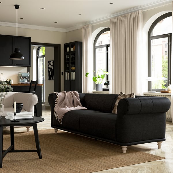Cozy living room with VISKAFORS black sofa, white curtains, and kitchen in background.