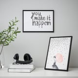 Framed posters on a desk. One says you make it happen, the other shows a woman with a red dot. Headphones on books, plant beside.