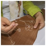 In handen zijn te zien die een bruine kussenhoes met witte bloemmotieven borduren. De stof is van katoen en het ontwerp heeft delicate handgestikte details.