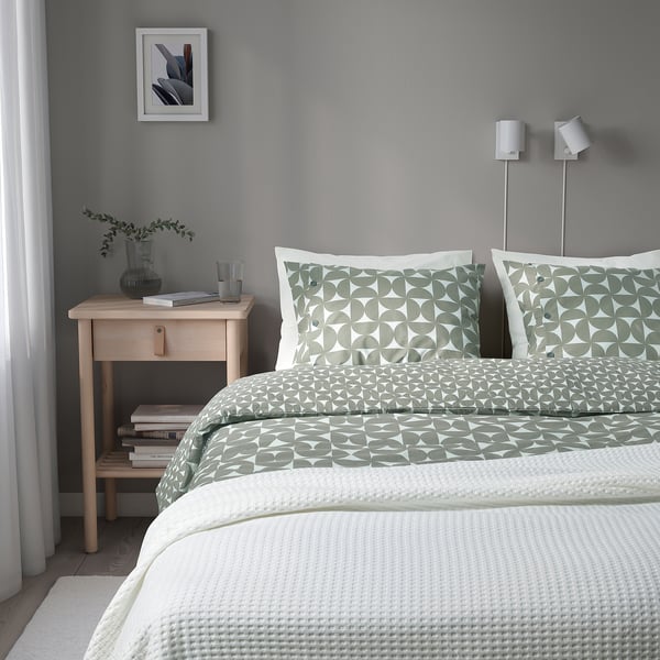 Nette slaapkamer met modern design: grijsgroen beddengoed met patroon, houten nachtkastje met boeken, glas en plant. Twee verstelbare zilveren lampen naast het bed.