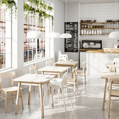 Modern café met witte tafels, stoelen en hanglampen; Grote ramen, weelderige planten, krijtmenubord en koffieapparatuur.