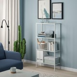 Moderne zilverkleurige metalen BAGGEBO plank met boeken en vazen tegen blauwe muur. Grote cactus en blauwe stoel in de buurt.