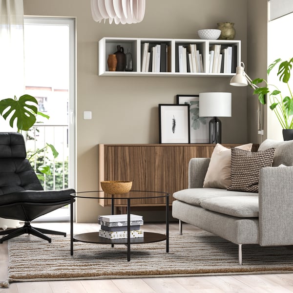 Moderne woonkamer met BANKKONTOR vloerkleed, grijze bank, salontafel, zwarte stoel, houtopslag en planten. De grootte van het vloerkleed is een aanvulling op meubels.