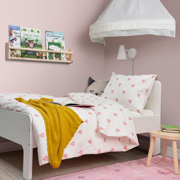 Klein kind wit bed met roze hart lakens, gele deken, kat hoofdkussen, muur boekenplank, lamp, kruk en kleed in een roze kamer.