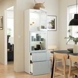 Een moderne, hoge kast met glazen deuren toont items terwijl andere in lades worden verborgen. Achter glas zijn grijze mokken en decoratieve voorwerpen zichtbaar.
