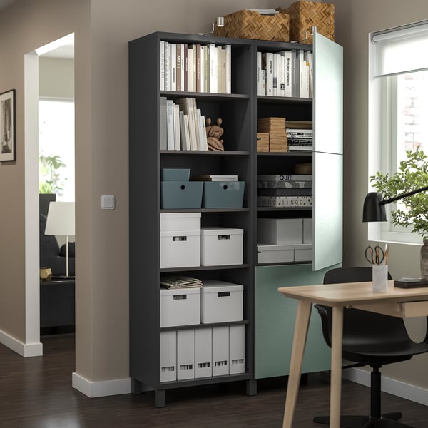 Modern kantoor aan huis met BESTÅ opbergmeubel in donkergrijs met open planken en kastdeuren, waarin boeken, dozen en decoratieve voorwerpen zijn ondergebracht.