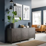 Moderne woonkamer met donkergrijs BESTÅ opbergmeubel, oranje bank, grote groene plant, hanglampen en ingelijste foto's aan een donkerblauwe muur.