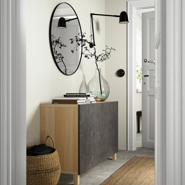 Moderne minimalistische entree met houten en grijze dressoircombinatie, grote ovale spiegel, zwarte wandlampen, potplant, boeken en een gevlochten mand eronder.