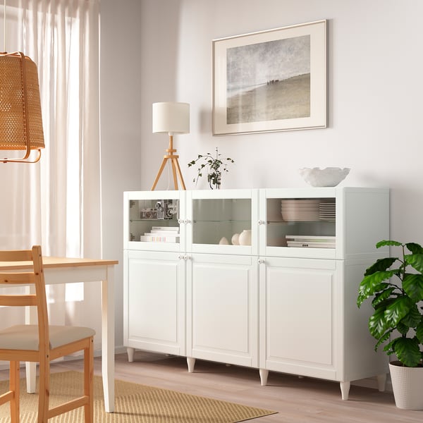 Modern BESTÅ dressoir met glazen deuren en lades, naast een houten stoel, met decor en boeken.
