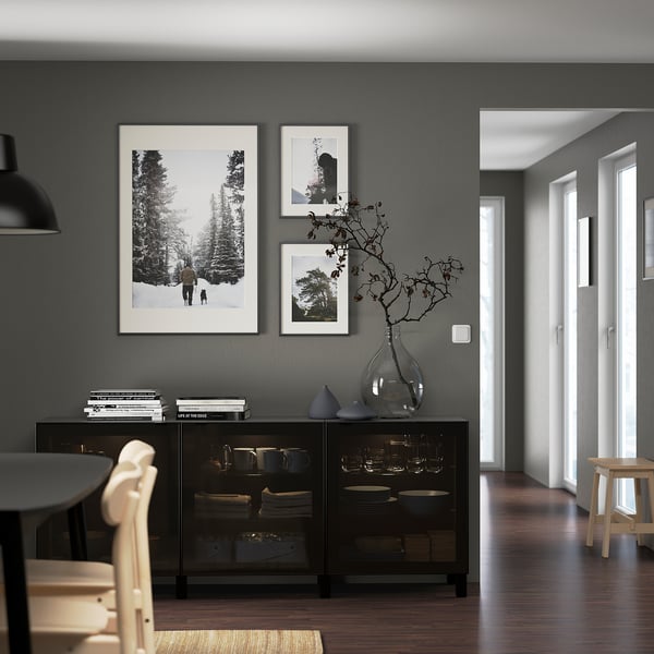 Moderne eetkamer met zwart BESTÅ dressoir, borden, takken, boeken, natuurfoto's, hanglamp en lichte stoelen rond zwarte tafel.