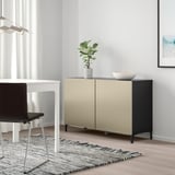Moderne kamer met beige en zwart BESTÅ dressoir, wit bureau, zwarte stoel, plant, kom en gestreept vloerkleed.
