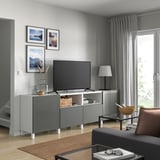 Moderne woonkamer met grijs BESTÅ tv-meubel, flatscreen-tv, grijze gordijnen, houten vloer, bruin vloerkleed, ingelijste foto's, lamp en zwarte bank.