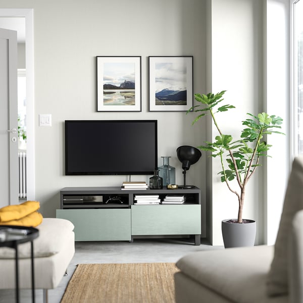 Moderne woonkamer met BESTÅ tv-meubel, flatscreen tv, decoratieve artikelen, boeken, gele deken en potplant. Lichtgrijze en mintgroene kleuren.