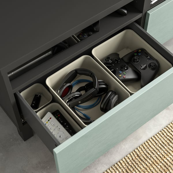 Open lade van grijs BESTÅ tv-meubel met compartimenten met gamecontrollers, headsets en afstandsbedieningen.