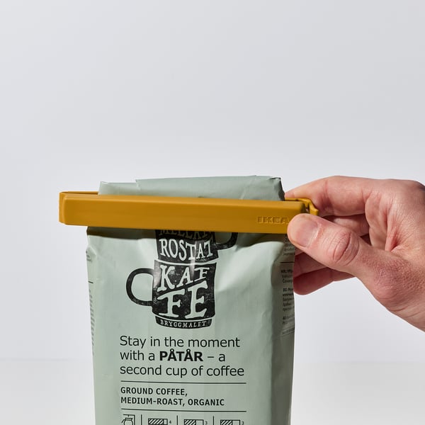 Hand sluit gele clip op koffiezak, verzegelt deze, zorgt voor versheid.