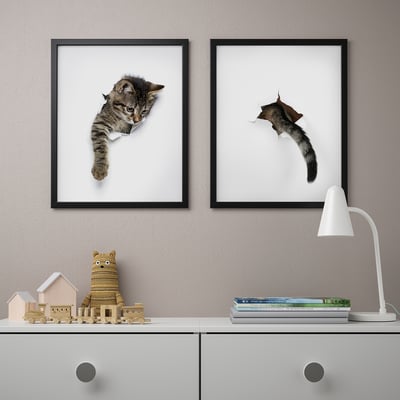 Boven een kast met snuisterijen en een lamp hangen twee fotolijstjes met een kattenthema.