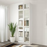 Een hoge witte BILLY boekenkast met glazen deuren, met boeken en decor, tegen een witte muur. Er staat een grote plant naast.