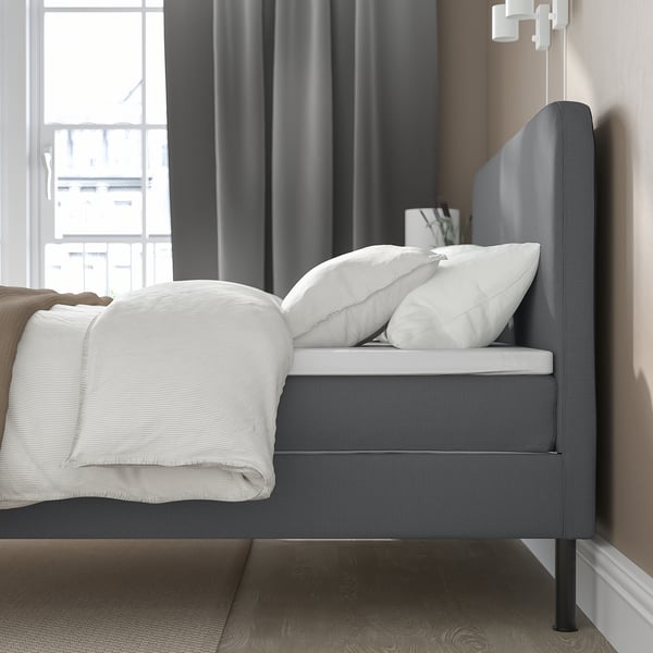 Modern bed met grijs gestoffeerd hoofdeinde, witte kussens en opgevouwen witte deken, geplaatst bij een groot raam met donkere gordijnen.