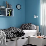 Modern bed met grijs geruit dekbed, wit frame, blauwe muur. Klok, boekenplank, bijzettafel met lamp en boeken.