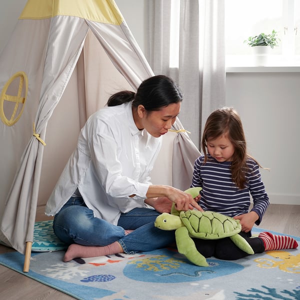 Vrouw en kind spelen met zacht groen schildpadspeelgoed in de BLÅVINGAD-tent.