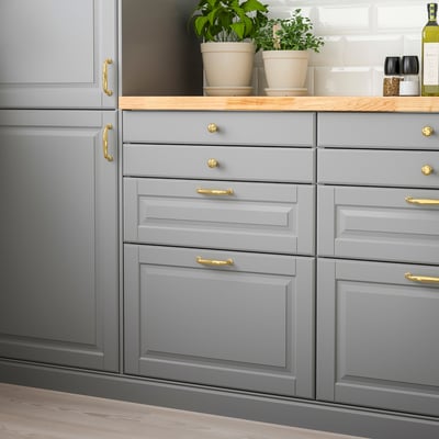 Een grijze keukenkast met BODBYN-design, met gladde, afgeschuinde panelen en gouden handgrepen, gepositioneerd onder groene planten en in de buurt van opbergartikelen.
