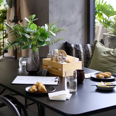 Zwarte tafel met kruikennpotten, groen, eten; houten kist met flessen; een heldere vaas met planten; borden en kopjes.