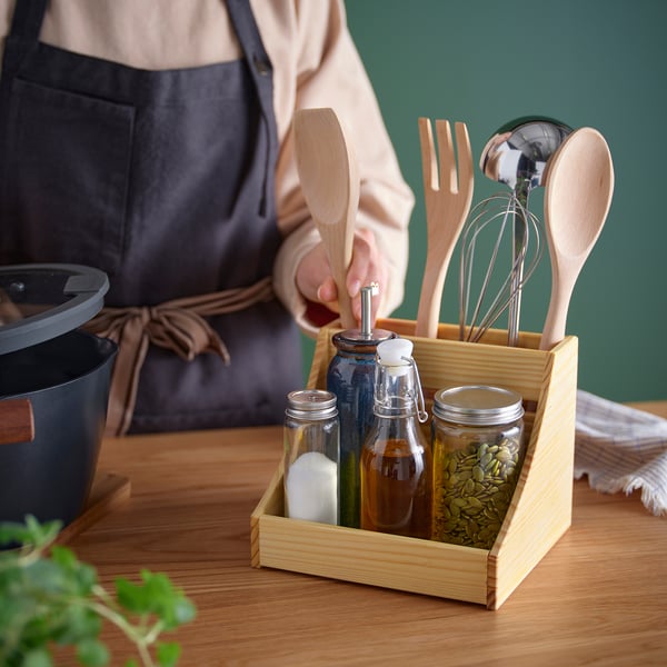 Toont een persoon die voedsel bereidt met een houten keukengereihouder met daarin verschillende houten kookgerei en kruidenpotten.