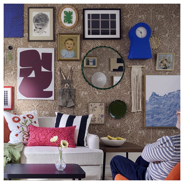 Levendige woonkamer met bloemenbehang, witte bank, eclectische kussens, galerijmuur en een persoon in gestreepte broek.