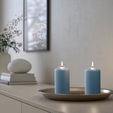 Twee brandende blauwe DAGLIGEN kaarsen op een dienblad, rechthoekige vorm, geplaatst op een plank. Vlakbij een stapel boeken en een witte vaas met bloemen.