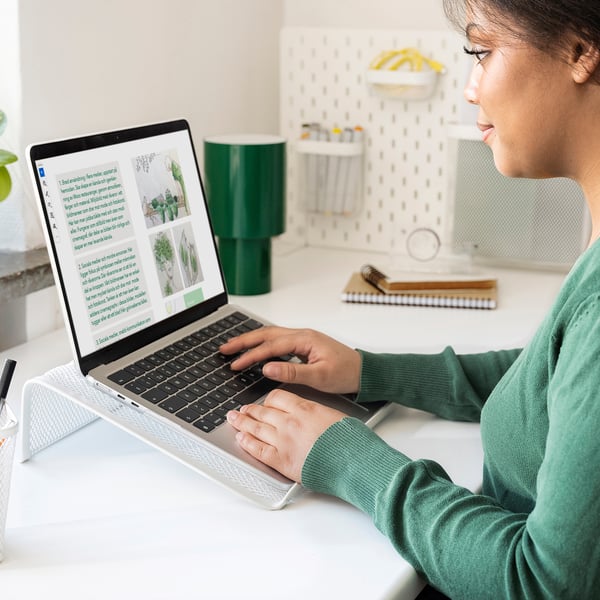 Met behulp van zilveren DRÖNJÖNS laptopstandaard op een wit bureau, met groene lamp, voor een betere houding.