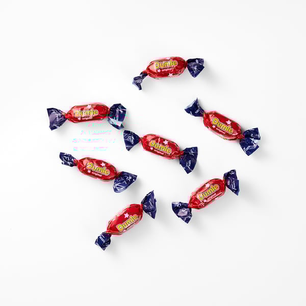 Acht rood verpakte DUMLE-snoepjes met blauwe uiteinden liggen verspreid over een wit oppervlak. DUMLE gladde chocolade en zachte taaie toffee vulling.