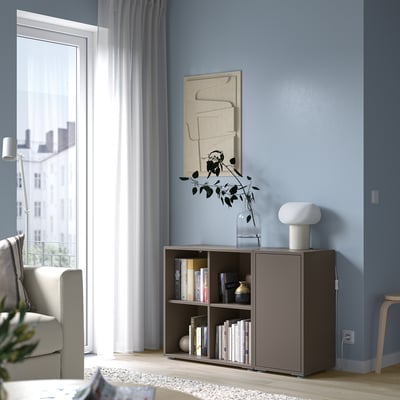 Moderne woonkamer met blauwe muur, abstracte kunst, boekenplank, stoel en raam met doorschijnende gordijnen. Focus op EKET opbergmeubel met boeken en vaas.