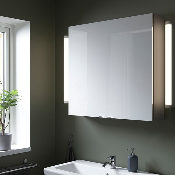 Moderne badkamer met grote glanzend witte ENHET kast, spiegeldeuren, verlichting. Donkergroene muren, gootsteen, plant en handdoek in de buurt.