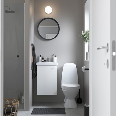 Moderne badkamer met witte ENHET kast, wastafel, toilet en ronde spiegel erboven. Compact en ruimtebesparend.