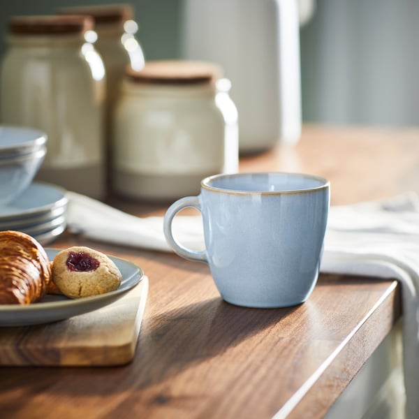 Een lichtblauwe FÄRGKLAR-mok staat op een houten tafel in de buurt van gebak en borden en toont het eenvoudige en functionele ontwerp in een gezellige omgeving.