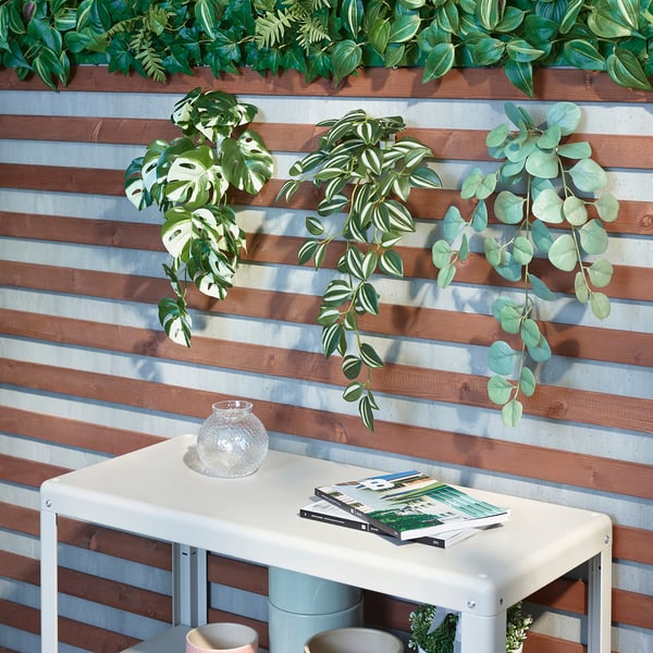 Aan een houten muur hangen drie kunstplanten, waardoor een groene, ontspannende ruimte ontstaat. Een witte tafel eronder bevat tijdschriften en een decoratieve vaas.