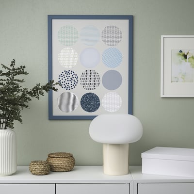 Kleurrijke cirkel art print op groene muur met lamp, mand, plant eronder; Nog een afdruk rechts.