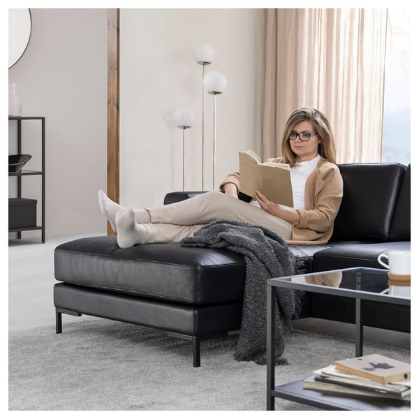 Vrouw leest op FULLERÖ zwarte leren bank, met een beige trui, een beige broek en sokken. De bank heeft strakke lijnen, zwarte poten en een grijze deken.