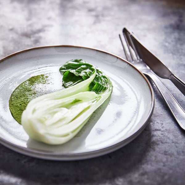 Een GLADELIG aardewerken bord, lichtgrijs met een zandglazuur, met een groene bladsalade. Een mes en vork staan ernaast op een donkere tafel.
