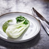 Een GLADELIG aardewerken bord, lichtgrijs met een zandglazuur, met een groene bladsalade. Een mes en vork staan ernaast op een donkere tafel.