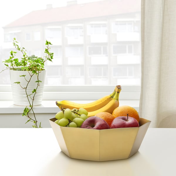 Een gouden GOKVÄLLÅ-schaal houdt fruit bij een raam. Het heeft de vorm van een achthoek en staat naast een potplant, wat het compacte maar elegante ontwerp benadrukt.