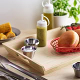 Houten serveerplank met handvatten, kleine metalen bekers, maïs, hamburgers in mand, kruiden, tang. GRILLTIDER-product .