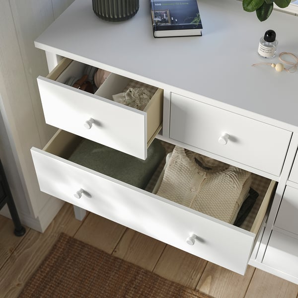 Wit dressoir met drie open lades die netjes opgevouwen kleding onthullen. Top heeft een boek, plant en andere items. Veiligheidsfunctie opgemerkt.