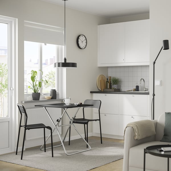 Moderne minimalistische kamer met witte muren, grijze accenten, opklapbare tafel en zwarte stoelen. Kleine keuken, groot raam, gezellige zitplaatsen.
