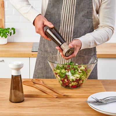 Een persoon maalt peper in een slakom met een HALVTOM kruidenmolen op een houten tafel.